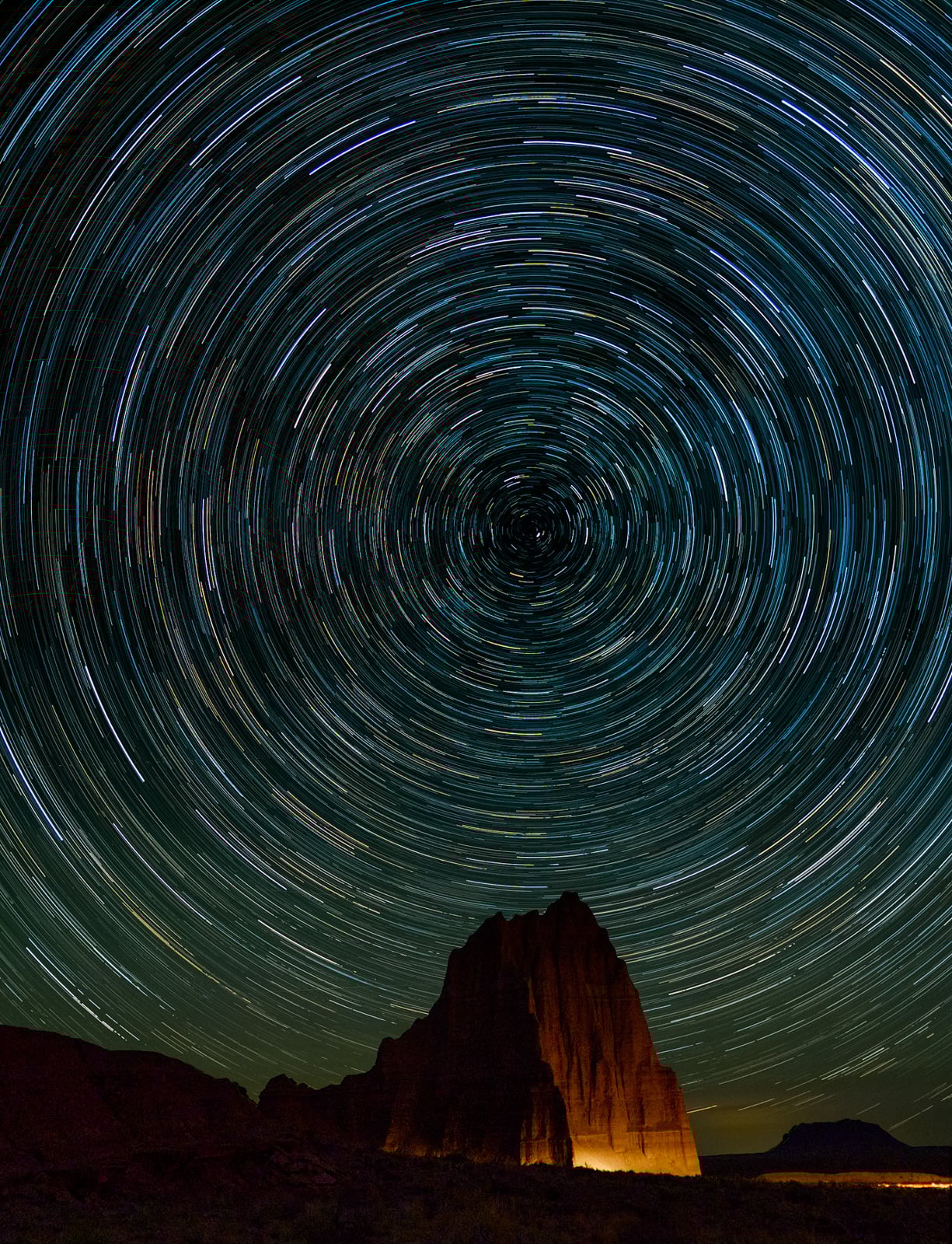 Long-exposure photo of circular star trails in the night sky above a dark mountain peak, with a faint warm light illuminating the base of the mountain.