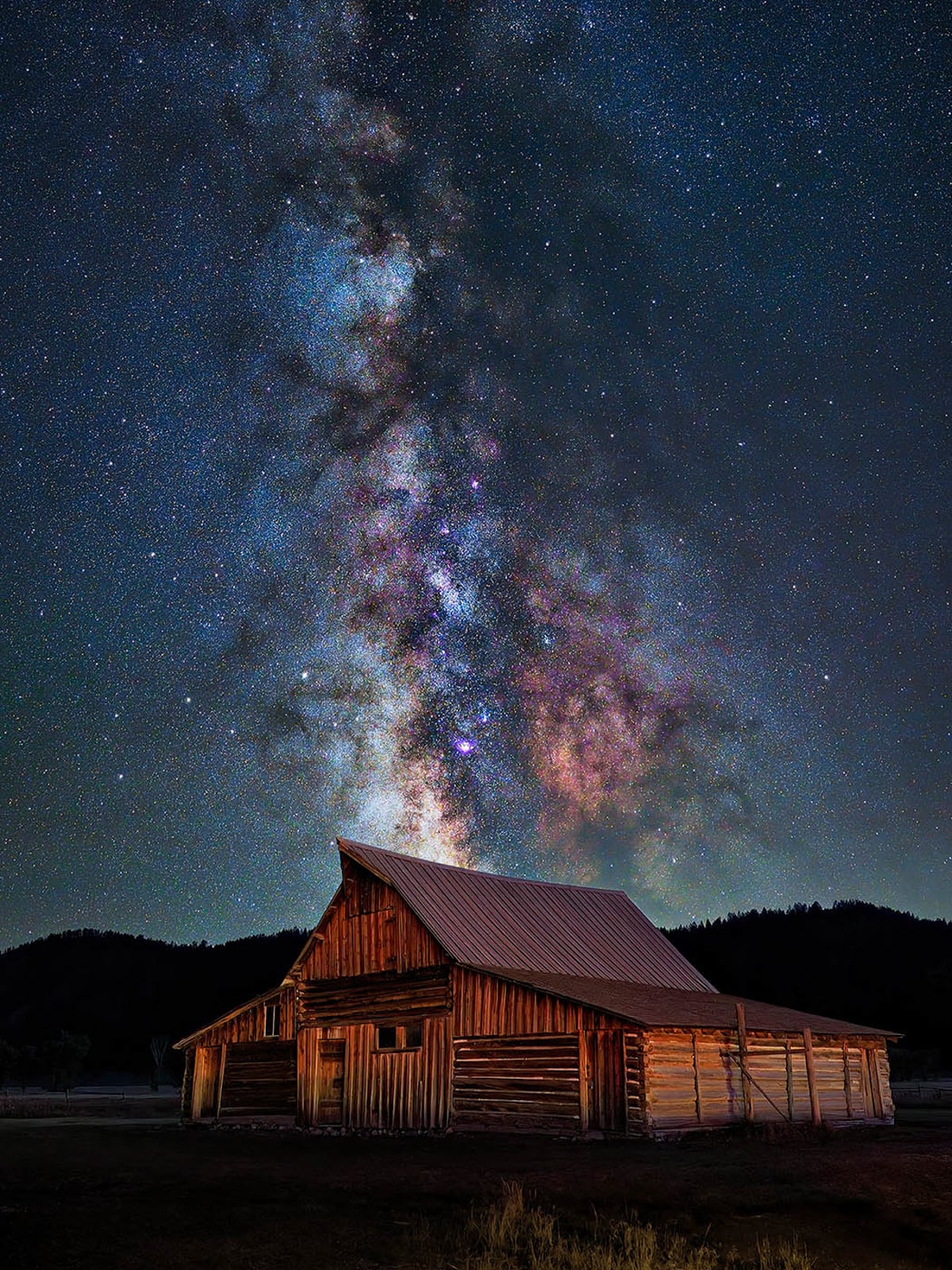 A rustic wooden barn stands in the foreground under a clear night sky, with the Milky Way’s stars and colorful nebulae vividly stretching upward above the roof.