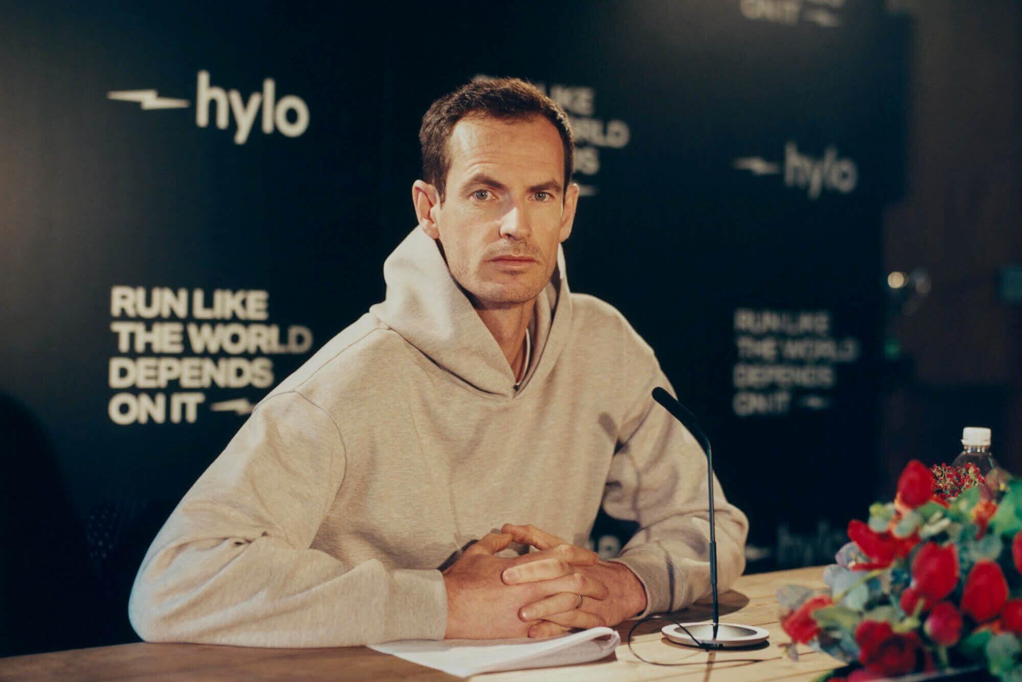 Andy Murray sits wearing a beige hoodie in front of a backdrop with a Hylo logo and slogan on it.