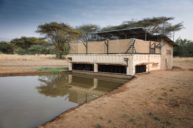 A beige shipping container structure with solar panels and shaded fencing sits beside a small pond in a dry, grassy landscape with scattered trees. The building's reflection is visible in the water.