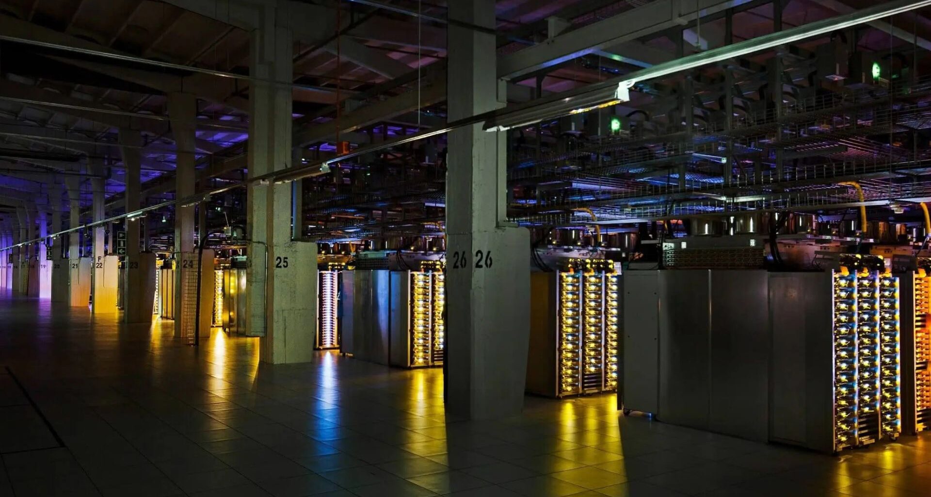 A large data center with rows of illuminated server racks and numbered concrete pillars under dim, colored lighting.