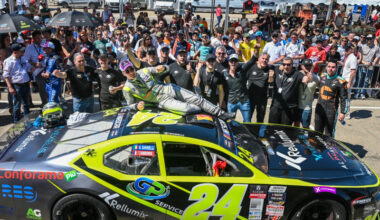 Yellow and black race car #24 with crowd of cheering fans and team members celebrating around it, one person lying on the car’s roof as fans wave and smile under tents.