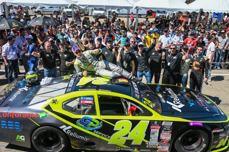 Yellow and black race car #24 with crowd of cheering fans and team members celebrating around it, one person lying on the car’s roof as fans wave and smile under tents.