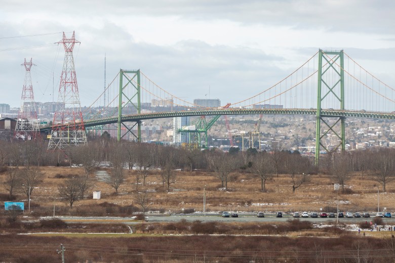On a grey winter day, the MacKay Bridge is seen in the background with the vacant lands known as Shannon Park in the foreground. On the right, there are parked cars and children playing in a field.