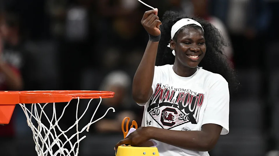 Madina Okoto, standing on a stepladder with a pair of scissors in her left hand, holds aloft a piece of basketball net which she has just cut off a basketball hoop next to her. She is wearing a predominantly white t-shirt and white headband, and is beaming with a smile