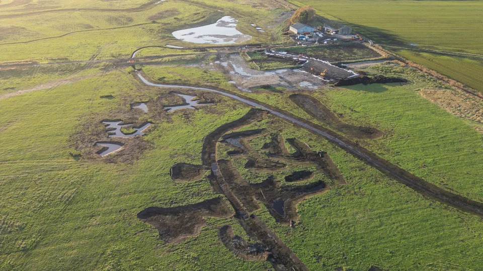 An aerial view of a flat landscape with a large pond and other rivulets nearby. Some agricultural buildings can be seen on the upper right of the image.
