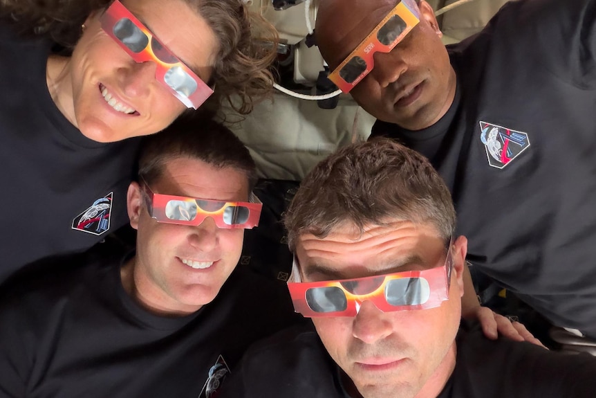 Three men and a women in navy shirts close together in zero gravity with cardboard eclipse glasses.
