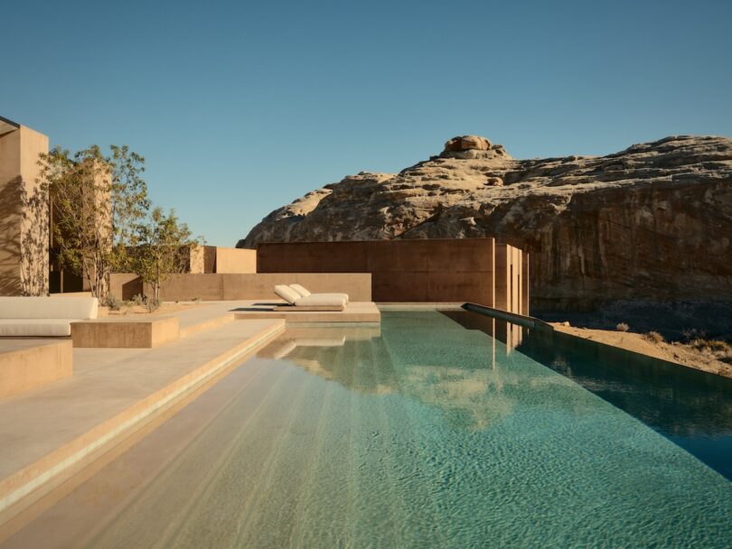 Modern infinity pool with lounge chairs, set against desert architecture and rocky landscape under a clear blue sky.