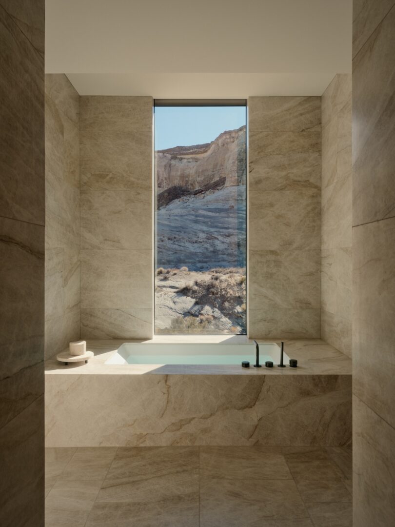 Modern bathroom with beige stone walls and floors, featuring a built-in tub below a tall window that frames a view of a rocky desert landscape.
