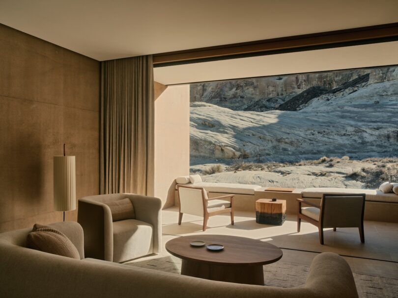 A modern, neutral-toned living room with large windows opens to a rocky desert landscape; minimal furniture includes armchairs, a sofa, and a round coffee table.