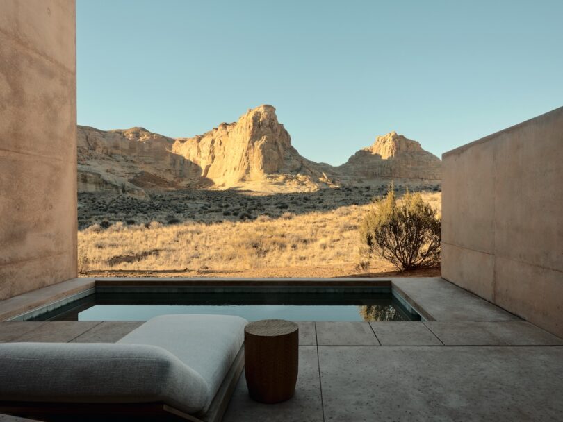 A small pool and lounge chair on a concrete patio overlook a sunlit desert landscape with rocky hills under a clear sky.