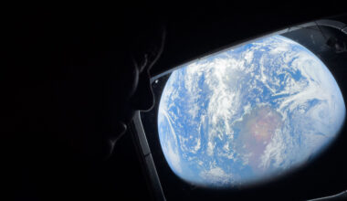 NASA astronaut Reid Wiseman looks out of the Orion spacecraft's rectangular window at Earth. He is barely visible in the dimly lit cabin, but Earth is bright. The entirety of Earth cannot be seen; in this view, Earth's round shape is cut off. Earth itself looks like a bright blue ball with swirling white clouds all over it; at center is a brown and green landmass.