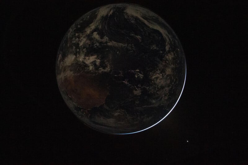 A view of Earth from space, mostly in shadow with a thin crescent of sunlight on the edge, and a small bright point of light visible against the black background.