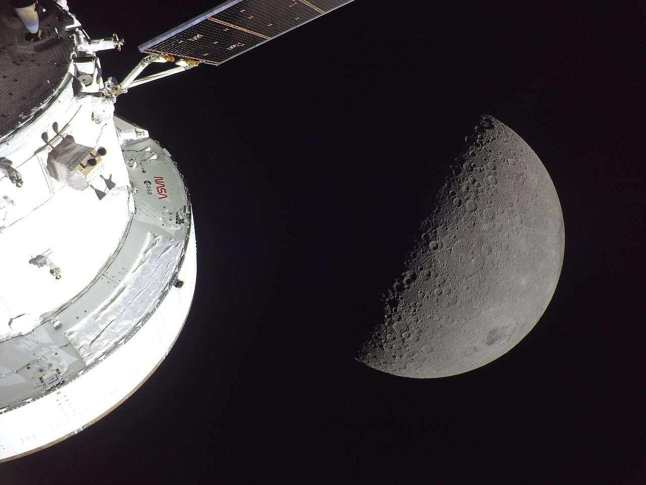 NASA’s Orion spacecraft is seen in the foreground, lit up by the Sun. A first quarter Moon is visible behind it, with sunlight coming from the right.