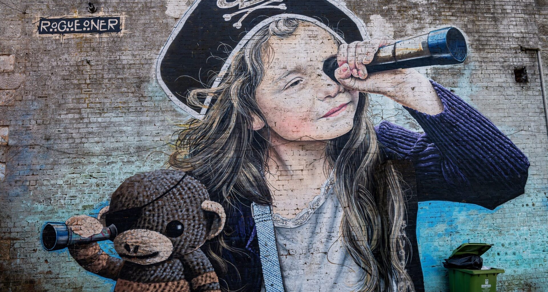 A mural by Rogue-one of a young girl with a pirate hat, looking through a spyglass, with her stuffed monkey toy also looking through a spyglass