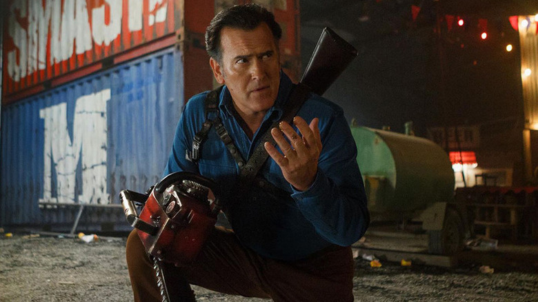 Ash Williams (Bruce Campbell) kneels by shipping containers with his chainsaw ready in Ash vs Evil Dead