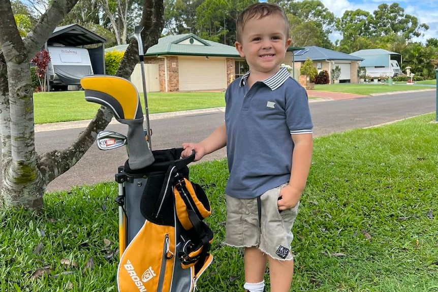 A young boys standing proudly next to his golf clubs and bag