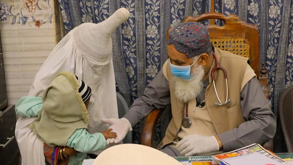 A man sitting in a chair wearing a stethoscope, face mask, and gloves greets a woman holding her child. The woman and child's faces are obscured.