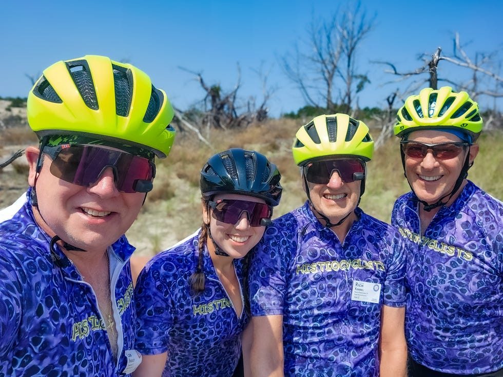 Four cyclists wearing helmets and matching jerseys, posing in a sunny outdoor setting. Four cyclists wearing helmets and matching jerseys, posing in a sunny outdoor setting.