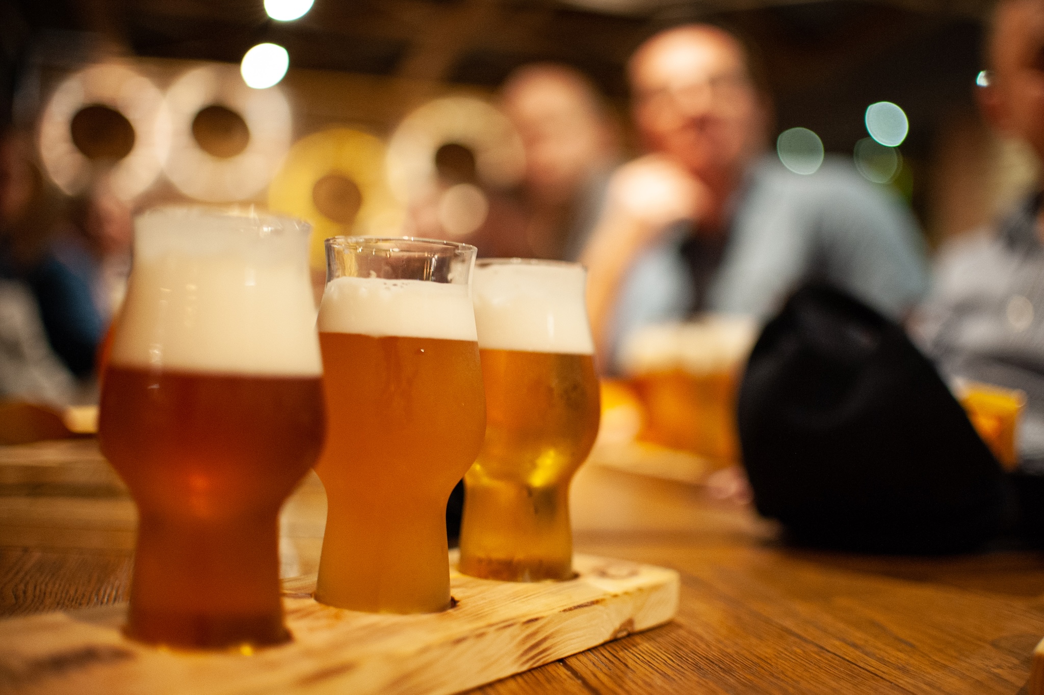 Three glasses of beer on a tasting tray with some blurred out people in the backgrodund.