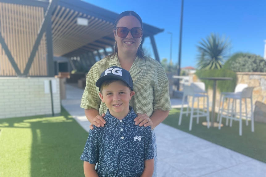 A young boy wearing a golf cap and polo shirt standing in front of a his mother 