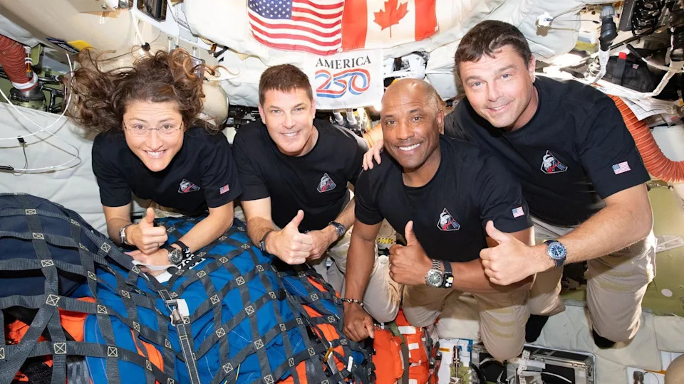 Four astronauts in dark blue T‑shirts float close together inside a bright, cluttered spacecraft. Their faces are blurred, but all give thumbs‑up to the camera. Their shirts carry a matching mission patch and small US flag on the sleeve. Behind them, American and Canadian flags hang above a red‑and‑blue “America 250” logo. Around them are white padded walls, orange pipes, cables and tightly netted blue and orange cargo bags, emphasising the cramped zero‑gravity cabin.