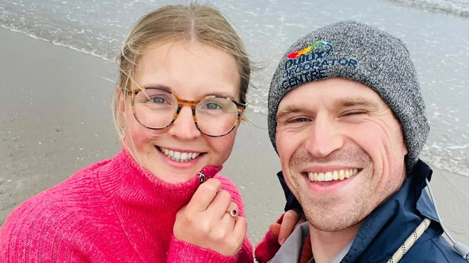 A young couple standing on a beach grin at the camera. The young woman, on the left, wears glasses and a bright pink jumper. On the right, the man wears a grey beanie.