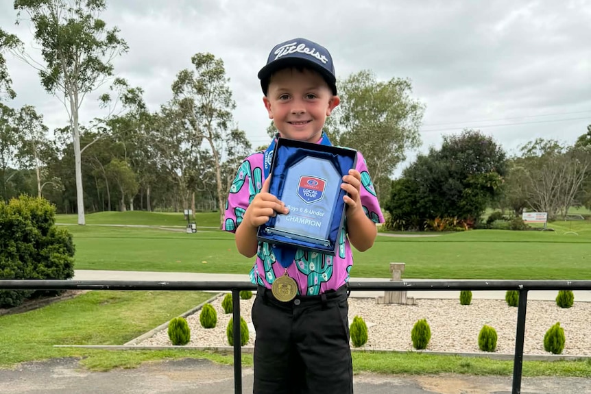 A young boy standing up wearing golf attire and holding up a plaque award 