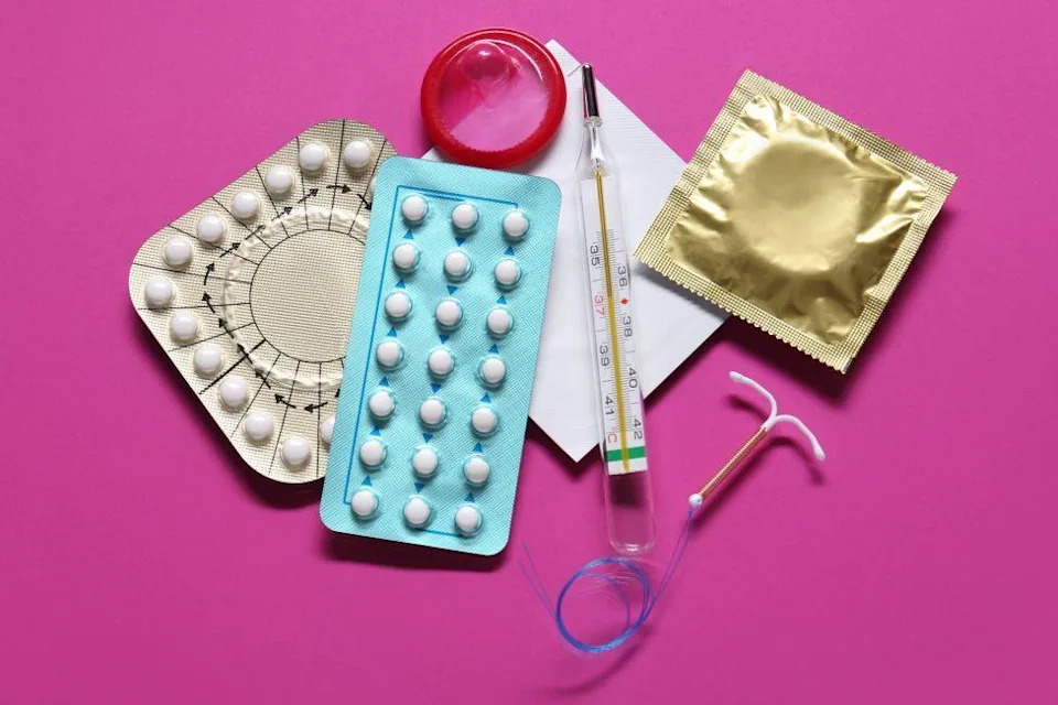 Photo of a range of contraception spread out on a table, including the pill, contraceptive coil and a condom in a wrapper, on a bright pink background