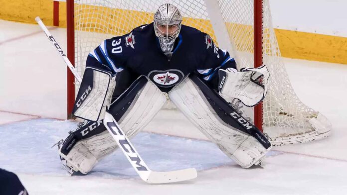 Connor Hellebuyck, star goaltender for the Winnipeg Jets, in focused butterfly stance in full navy blue and white gear in front of the net during an NHL game, amidst recent trade rumors.