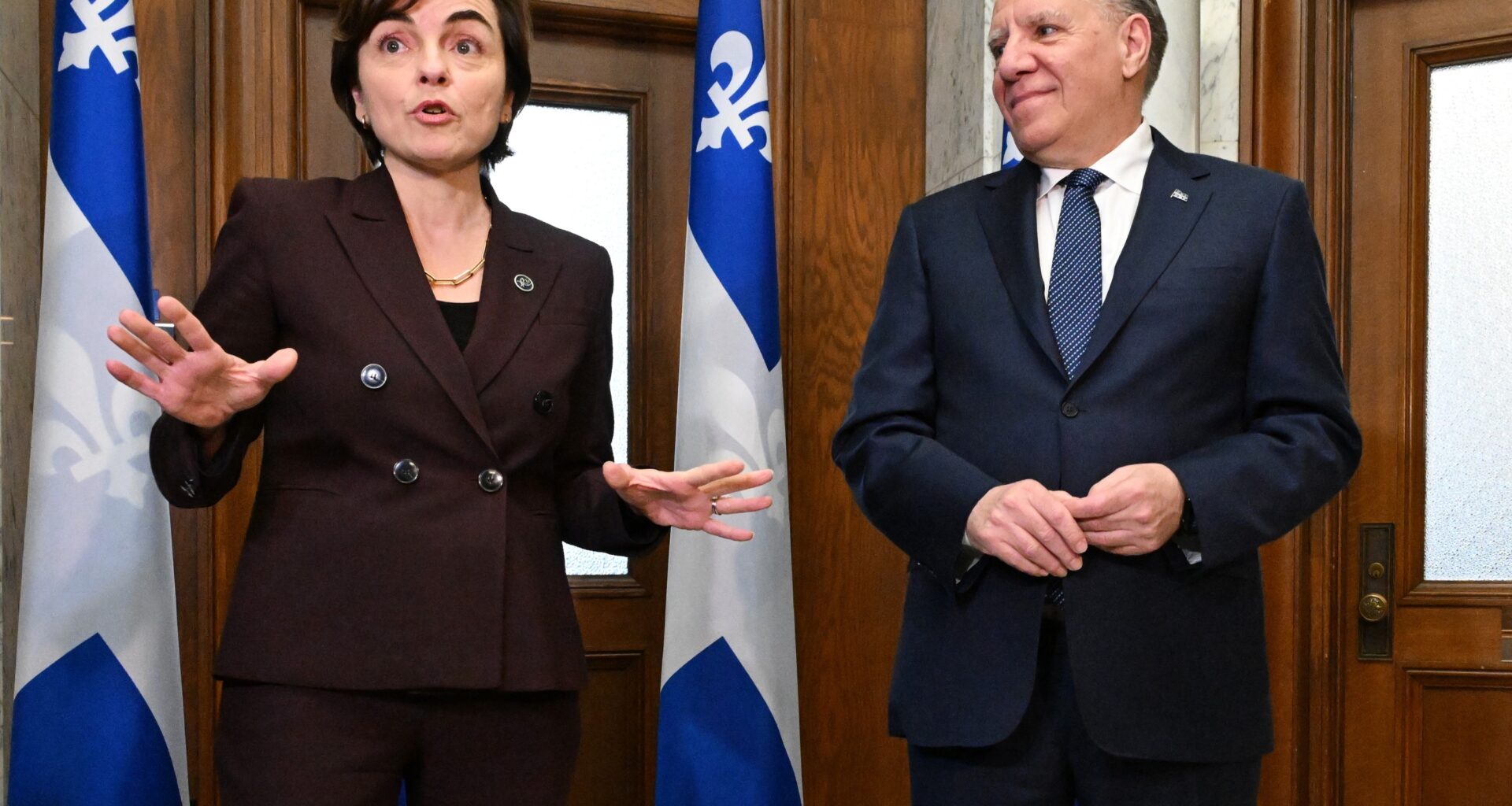 Christine Fréchette and François Legault stand and speak in front of doors and Quebec flags.