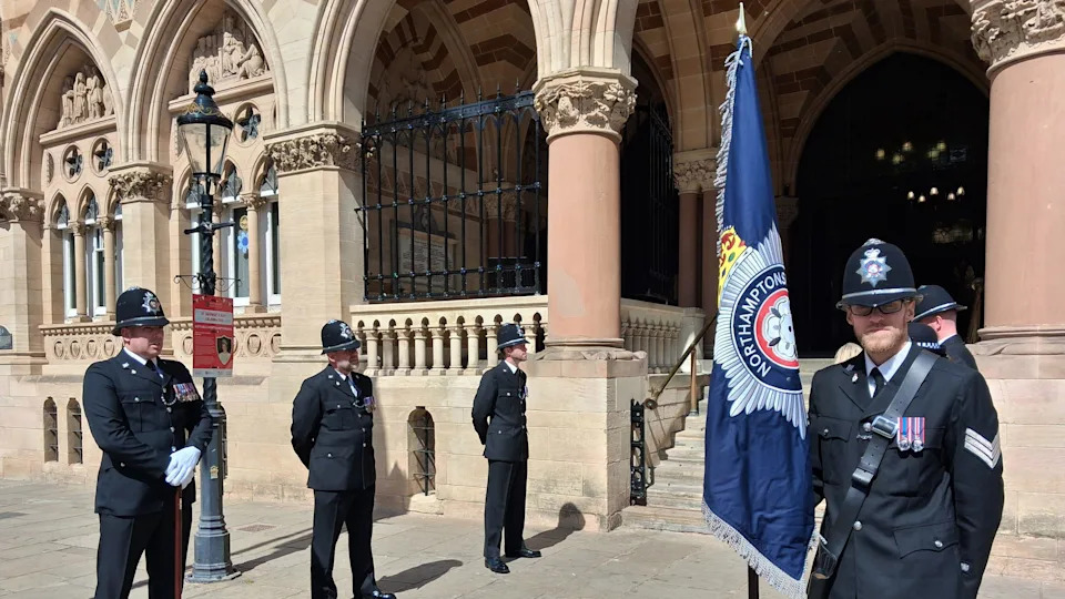 A police guard of honour stand in two lines facing each other outside a large civic building on a sunny day. One officer stands with a large blue flag with a police emblem on it and looks at the camera. All the officers are wearing matching uniform with black hats, black suits and trousers and white gloves. Some stand with their hands behind their backs while another to the left of the image has his hands in front of him as he holds a brown staff.