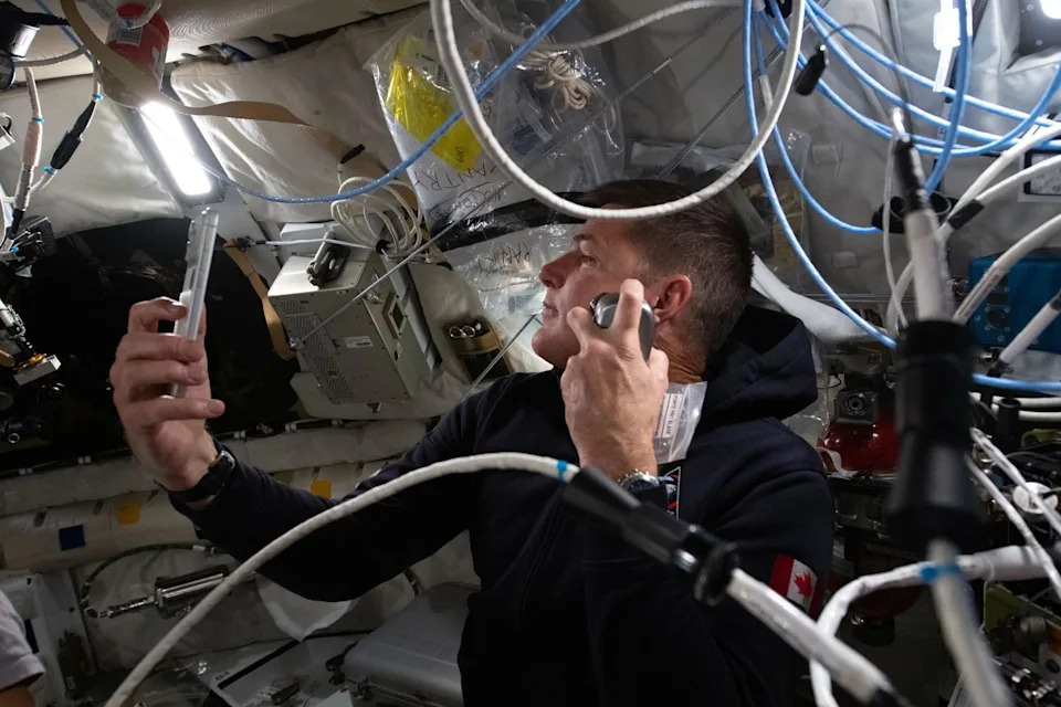 This close interior image shows Canadian astronaut Jeremy Hansen floating inside the cramped Orion spacecraft while shaving. He wears a dark hooded top with a small Canadian flag patch on the sleeve and holds a razor up to his cheek. Around him, the cabin walls are lined with white padded panels, storage bags and equipment boxes. Blue and white cables loop across the frame, and bright strip lighting overhead makes the small space look crowded, practical and lived in.