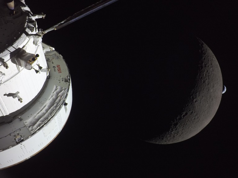 Part of a round, white spacecraft in front of the Moon, large, grey and partially in shadow, with the tiny crescent Earth visible in the background.