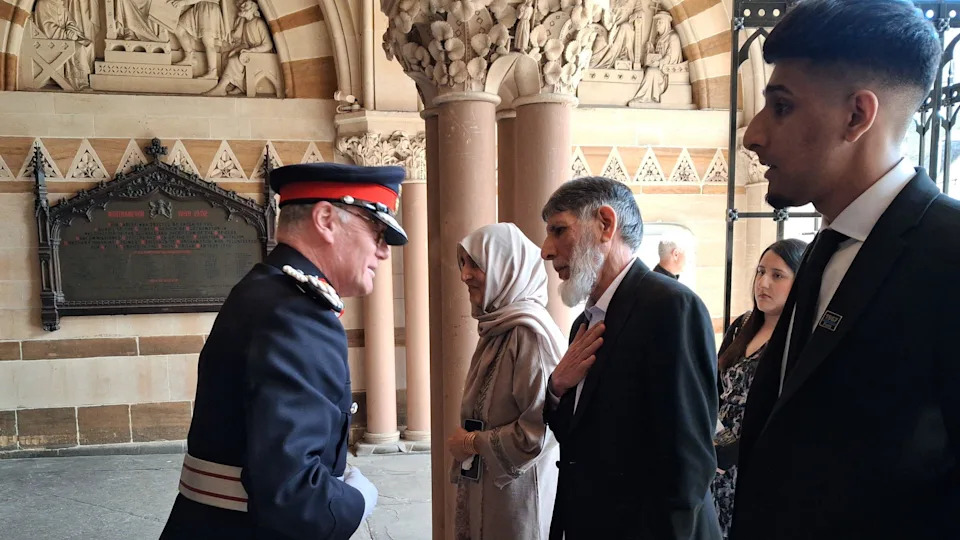 A police official wearing a hat and a police uniform greets members of a family at the entrance of a large ornate civic building. The officer speaks to a man in front of him who rests one hand on his chest. Other members of the family stand alongside him.
