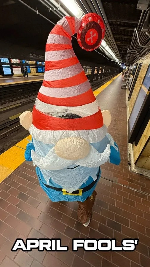 Person wearing an inflatable gnome costume with a tall red and white hat stands on a subway platform. Text reads "April Fools'"