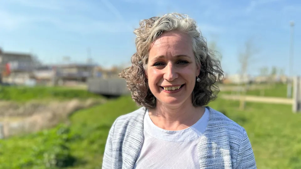 Rebecca Britton smiling into camera with green space and a housing development in the background. She has curly grey hair and is wearing a grey cardigan and white t-shirt.