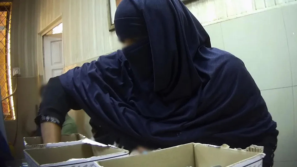 A woman puts her hands inside a cardboard box - two other boxes are visible. She is wearing a head and face covering, and her eyes are blurred for anonymity.