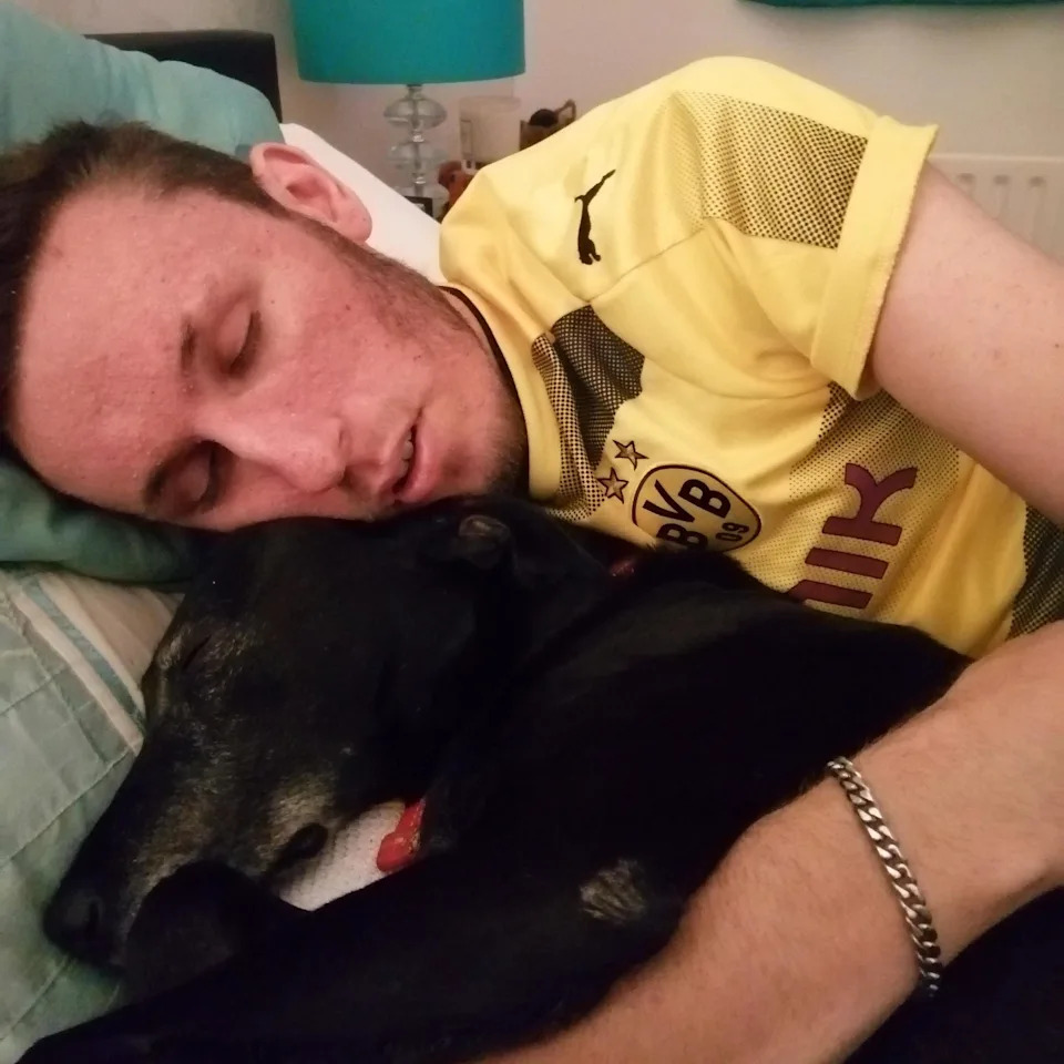 A young man on his bed asleep, cuddling his dog. The man is wearing a yellow football shirt and has his left arm over the dog. The dog has black fur.