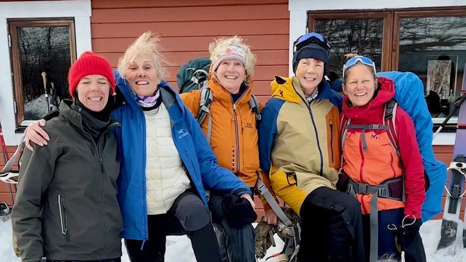 Five women in ski gear smiling to camera hugging each other