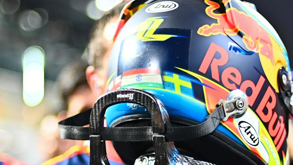 The back of Arvid Lindblad's F1 helmet. It shows the three flags of England, white with a red cross, India's horizontal lines of orange, white and green with a blue circle in the middle then on the right, Sweden's blue background with a yellow cross. The helmet also features the Red Bull sponsor with a red bull and yellow background with the writing of the brand below it. You can see the top of Arvid's racing suit and a black support for protecting his neck in a crash. 
