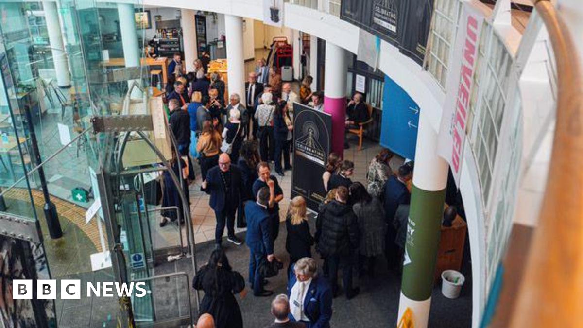 A crowd of people can be seen from above standing inside a building. There is a door to the left and columns through the middle. A large black sign is in front of the doors.