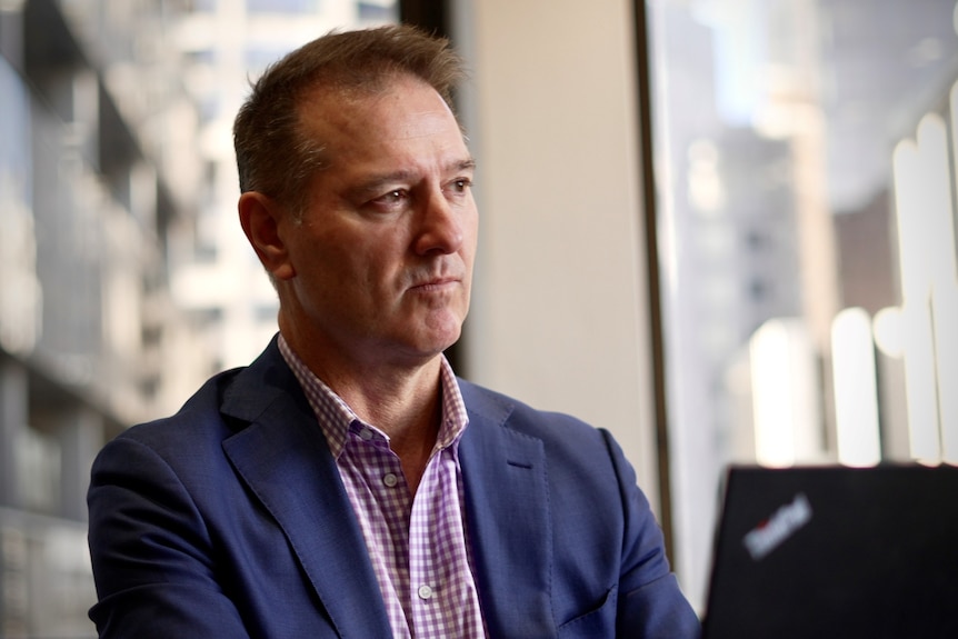 A middle aged man wearing a navy suit jacket and a purple and white checkered shirt.