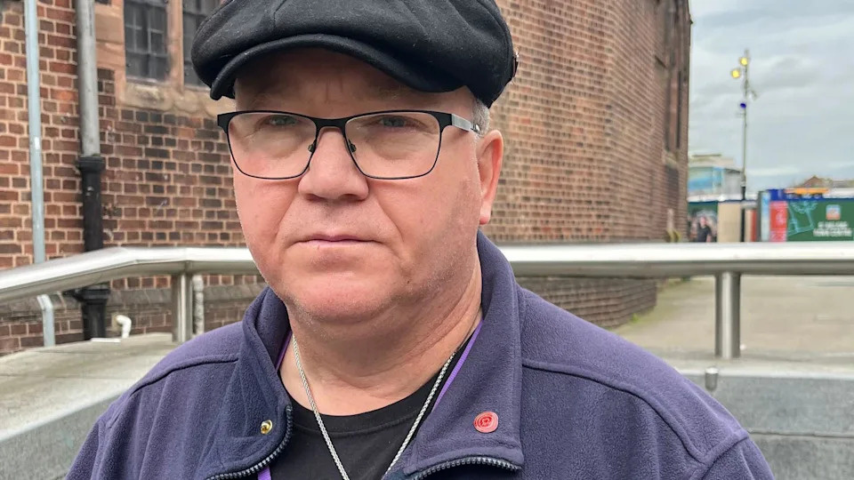 Graham Rudge, who has glasses, is wearing a black flat-cap and a purple fleece, looks into the camera with a serious expression.