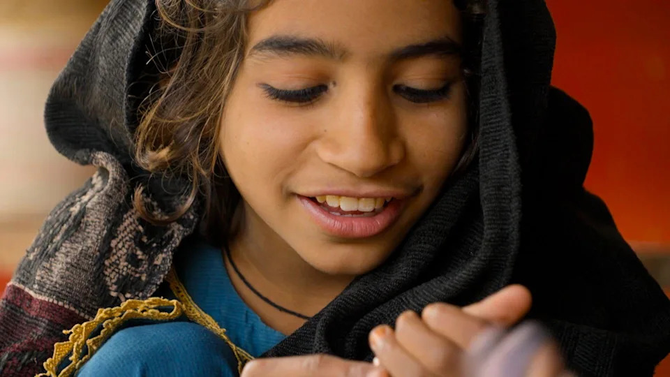 A young girl in a blue dress smiles, her eyes looking down towards her hands. She has a dark scarf with a patterned border over her head.
