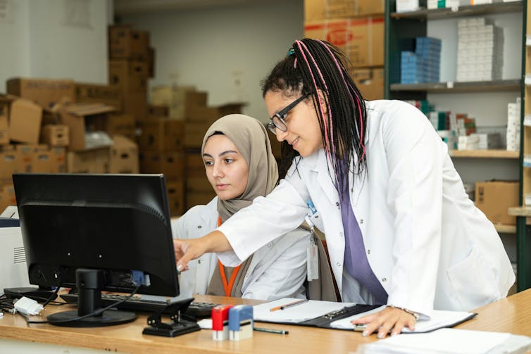 A woman points at a computer screen over the shoulder of a young woman, who is seated at a desk
