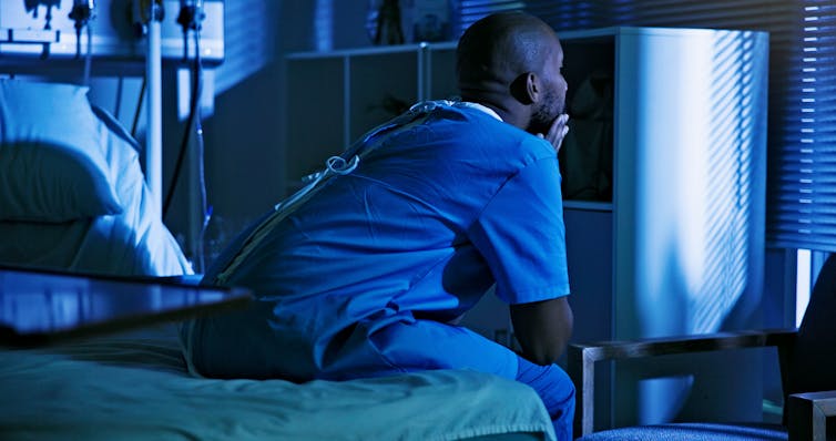 Patient in gown sitting on the edge of a hospital bed at night, elbows on knees and chin on clasped hands