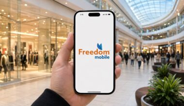 Close-up of a hand holding a smartphone displaying the Freedom Mobile logo in a bright, busy shopping mall with glass ceiling.