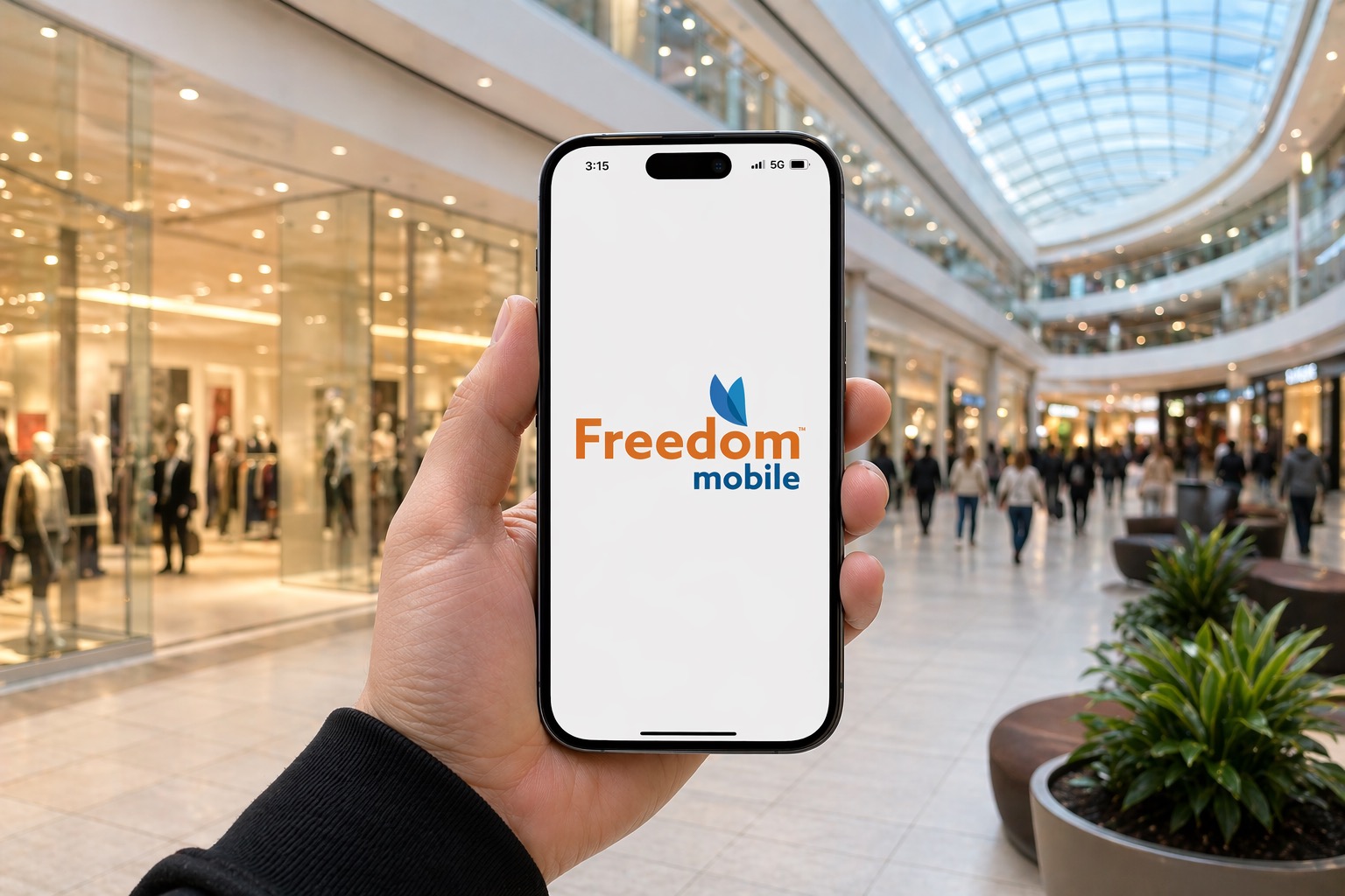 Close-up of a hand holding a smartphone displaying the Freedom Mobile logo in a bright, busy shopping mall with glass ceiling.