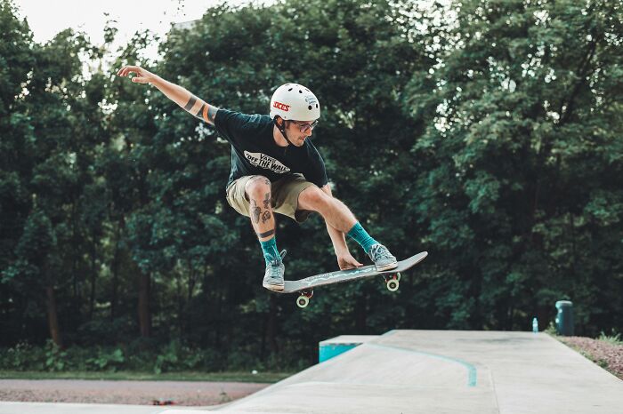 A person wearing a helmet performing a skateboarding trick at a skatepark surrounded by trees and greenery.